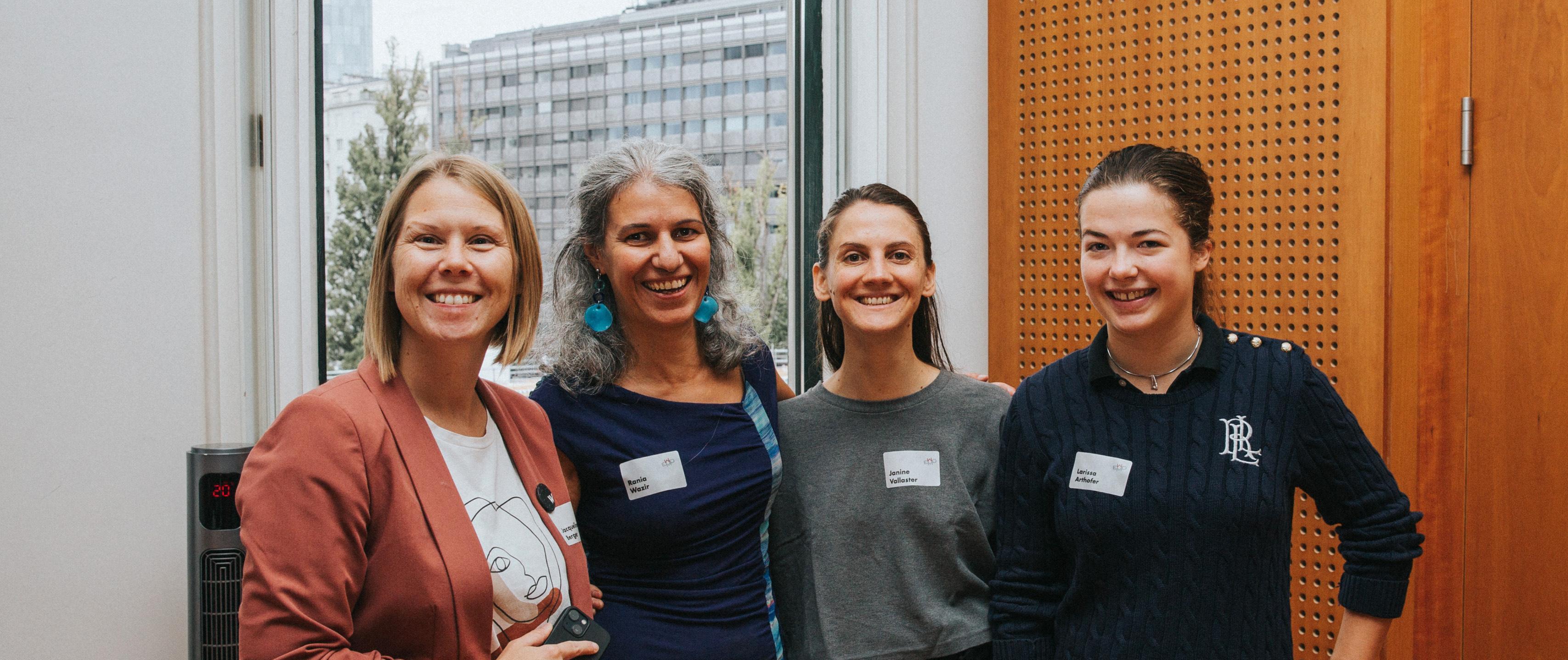 Zu sehen sind vier Frauen, die gemeinsam in die Kamera lächeln. Im Hintergrund ist ein großes Fenster zu sehen und auf der rechten Seite große Holz-Paneele. © Kathrin Hanga  - Women in AI Austria 